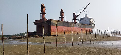 A large rusted ship at a shipbreaking yard with cranes on its deck